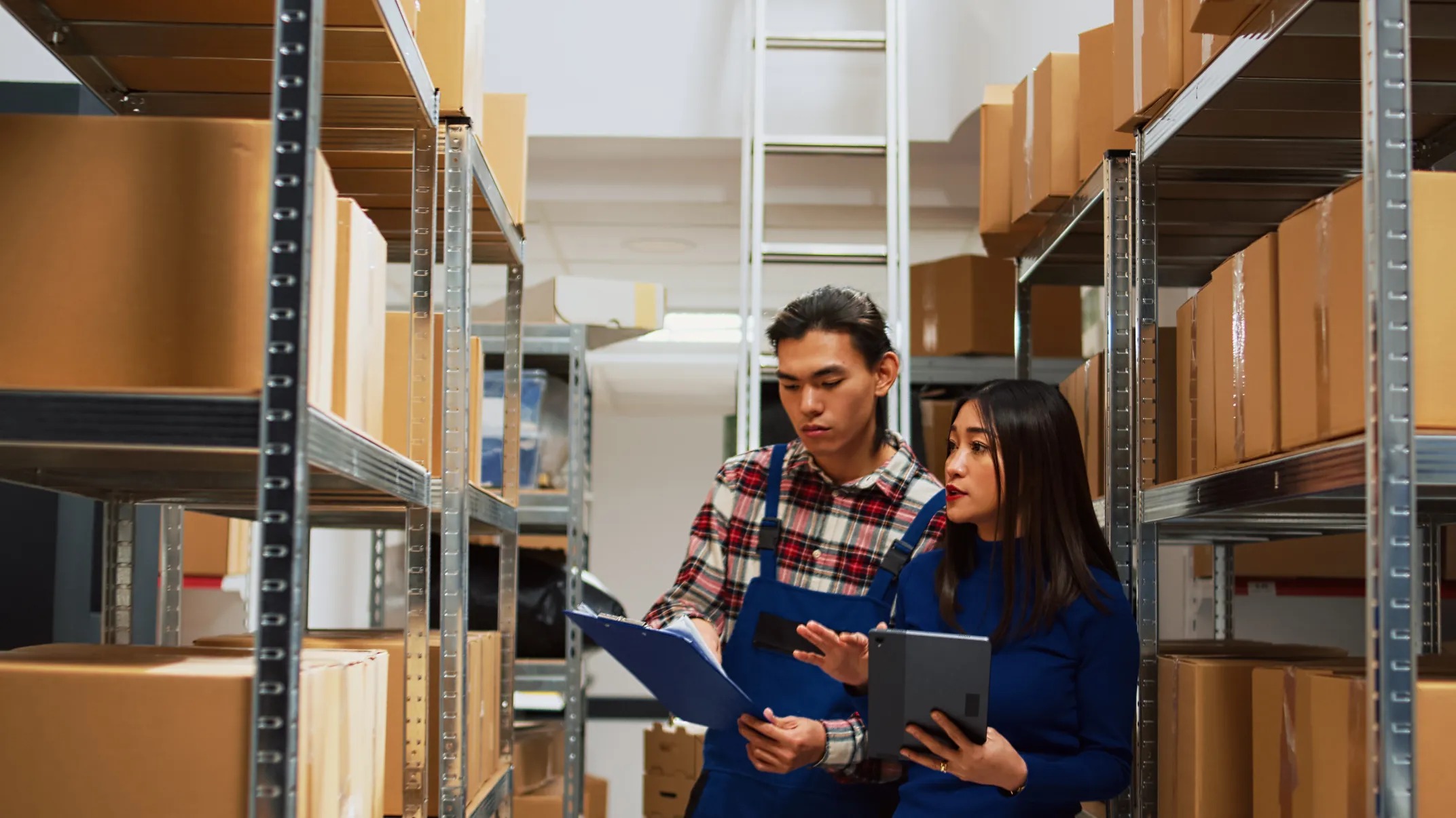 Two warehouse employees reviewing inventory on a clipboard and tablet between metal shelving units filled with cardboard boxes