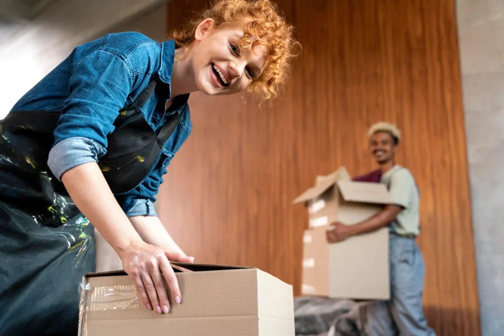 Cheerful woman with curly red hair and denim shirt leans over a cardboard box