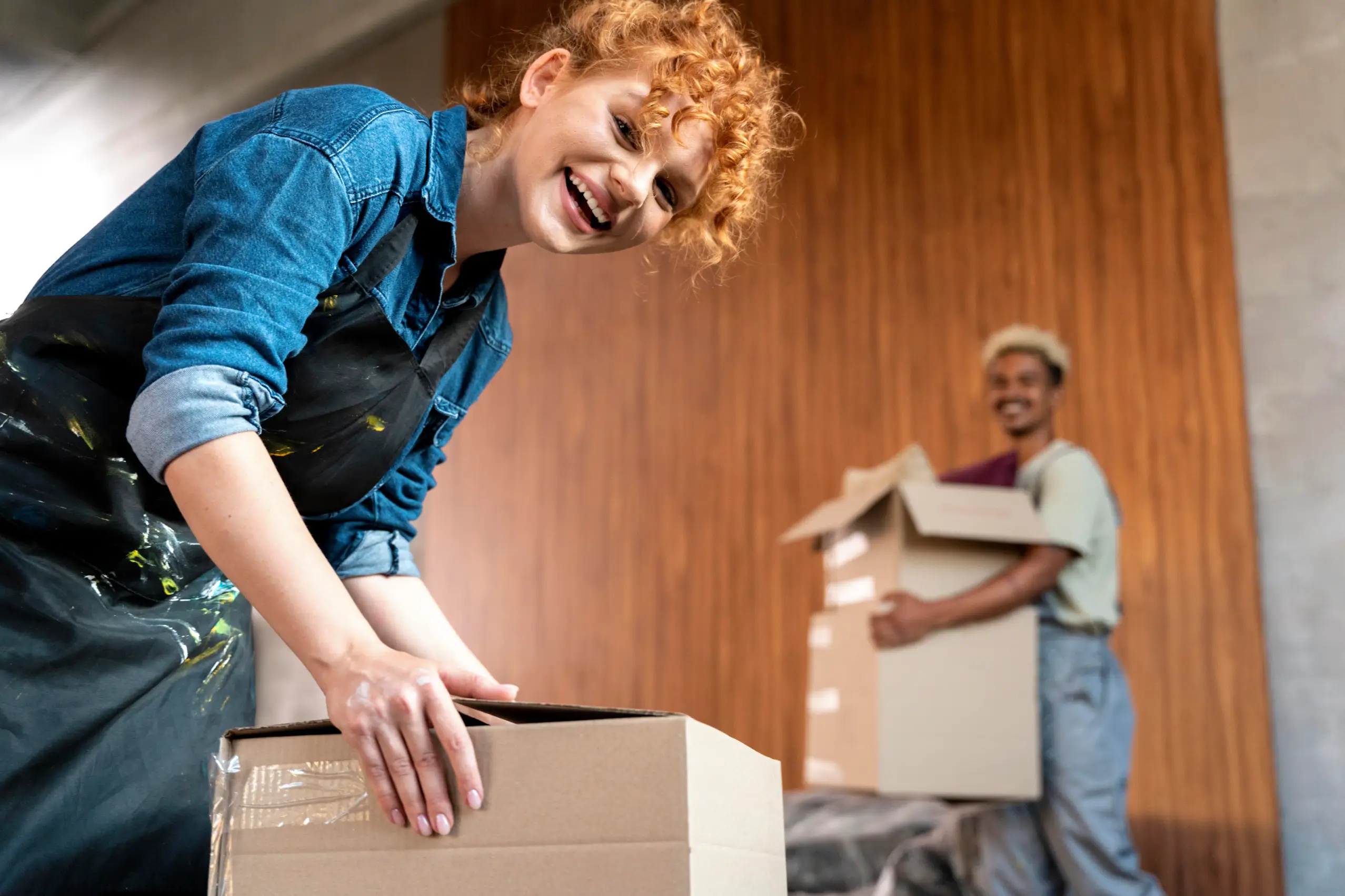 Cheerful woman with curly red hair and denim shirt leans over a cardboard box