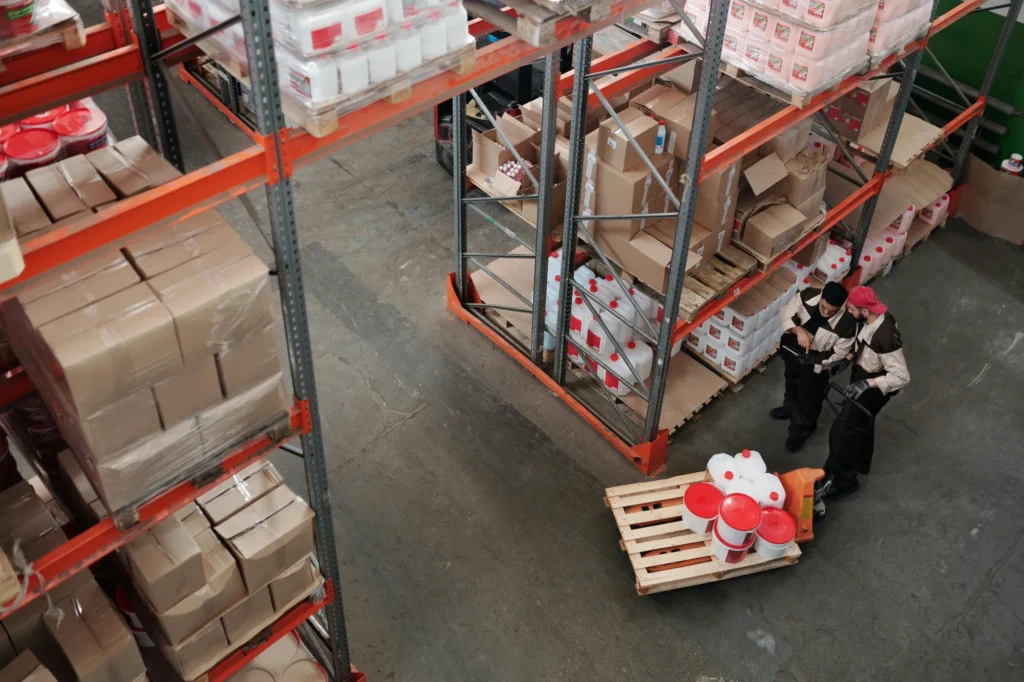 High-angle view of warehouse shelves stocked with boxes and containers, with two workers standing beside a pallet of large plastic jugs