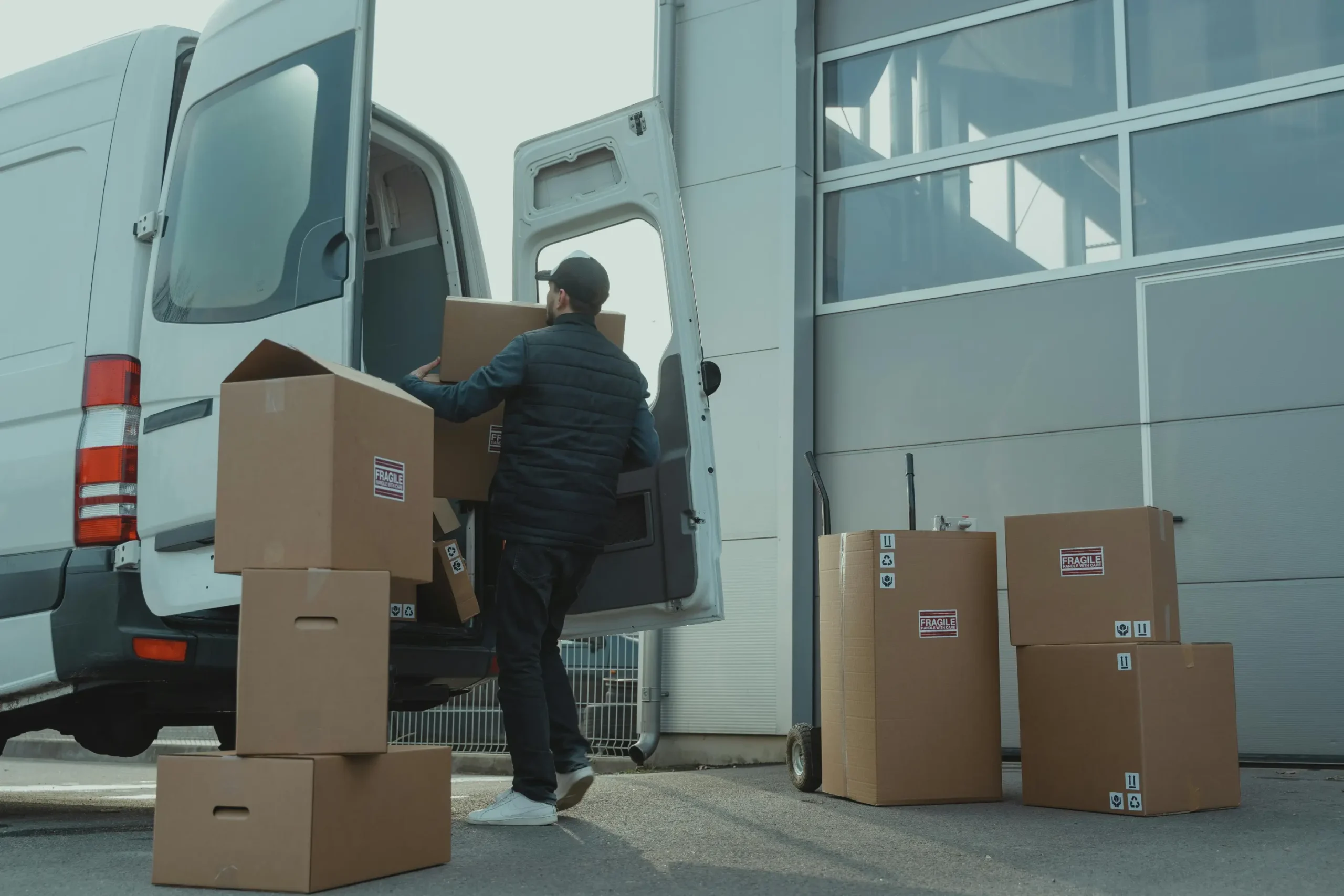 Delivery worker loading cardboard boxes labeled “fragile” into a white van outside a warehouse