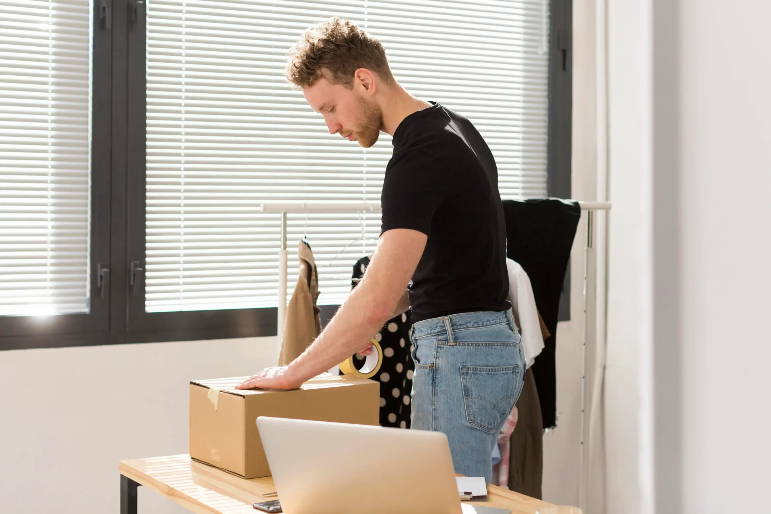 Young man wearing a black t-shirt and jeans seals a cardboard box with packing tape on a wooden desk