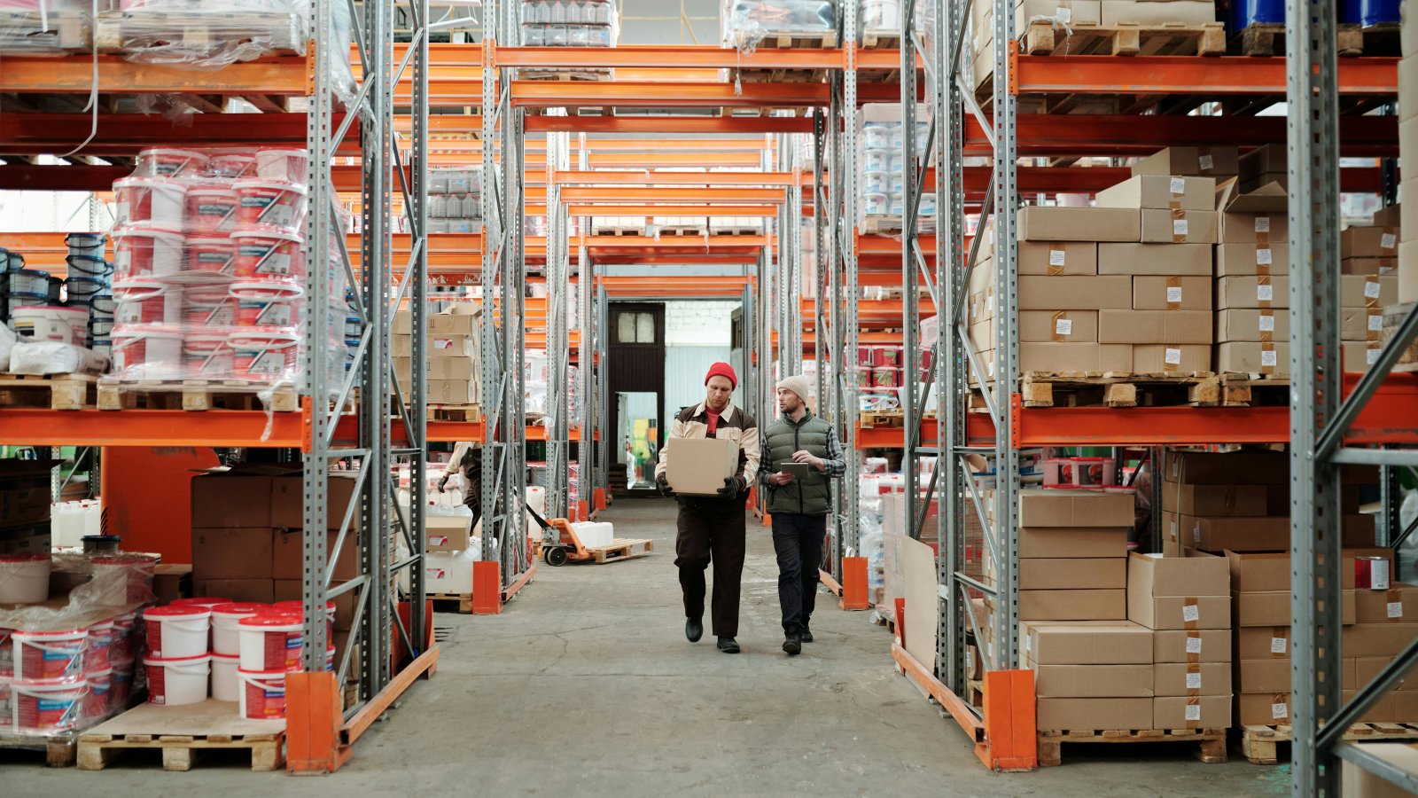 Two workers handle a package in a spacious warehouse with shelves stocked with boxes, representing warehouse logistics.
